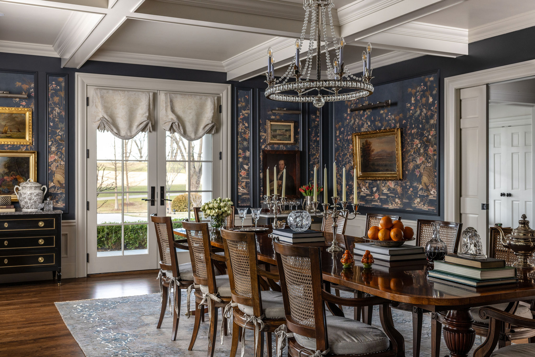 Wider view of Dining Room. The walls feature White wainscoting and a wallpapered navy blue, gold and oragne pattern with trim details by Rosette. These walls are adorned with gold framed artwork. Beautiful coffered ceiling in white with a crystal chandelier. Dark blue walls and a large bay window beyond a gorgeous wooden dining room table with oranges, large floral centerpieces and some candelabras.